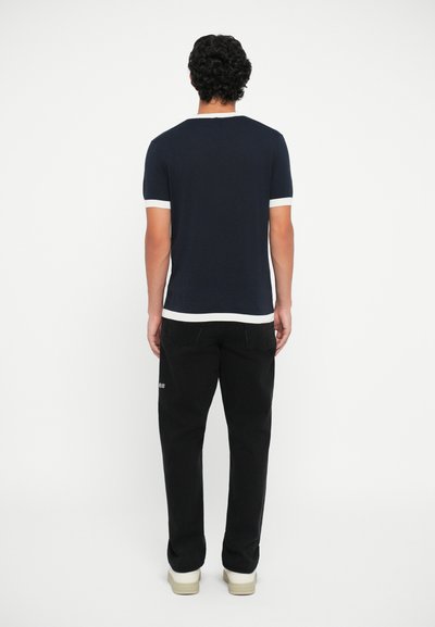 T-shirt de manga curta azul-marinho com acabamento branco no pescoço e nas mangas, combinado com calças pretas e sapatos de cor clara, visto por trás.