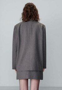 Blazer grigio a righe larghe con silhouette strutturata, maniche lunghe e orlo dritto, indossato sopra una gonna coordinata.