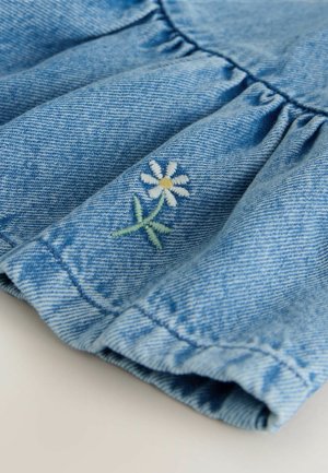 Denimová látka světle modré barvy s nařasenými volánky. Ve spodní části je vyšívaná bílá kopretina se žlutým středem a zeleným stonkem.