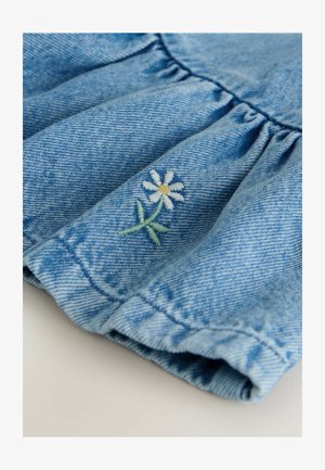 Traper tkanina u svijetloplavoj boji s nabranim naborima. Na dnu je ušivena bijela tratinčica sa žutim središtem i zelenim stabljikom.