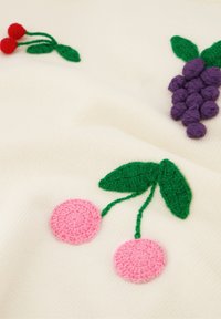 Witte gebreide stof met kleurrijke geborduurde fruit: rode kersen, paarse druiven en groene bladeren met textuurdetails.