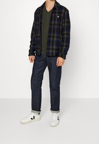 Abbigliamento maschile con camicia di flanella a quadri blu scuro e giallo, t-shirt a scollo a V verde oliva, jeans scuri e sneakers bianche con dettagli neri.