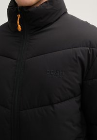 Sort polstret jakke med høj krave, der har en frontlynlås og tonale logo detaljer. Fremhævelser inkluderer en justerbar orange snørebånd.