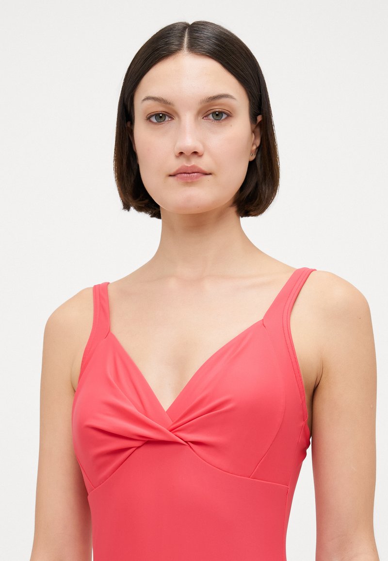 Maillot de bain corail rose avec un détail noué à l'avant, des bretelles fines et un tissu d'une texture lisse. Conception ajustée et décolleté angulé.