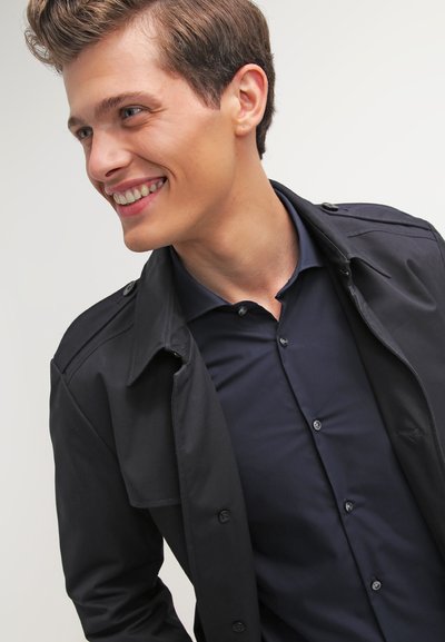 Un trench-coat noir sur une chemise boutonnée bleu marine, avec un col et des boutons à l'avant, en tissu lisse et au design ajusté.