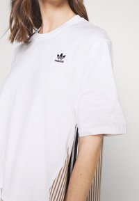 Valkoinen puuvillateeppari lyhyillä hihoilla, jossa on musta Adidas-logo ja raidalliset kultaiset ja mustat sivupaneelit.