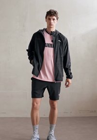 Svart zip-up jakke med subtil tekstur, rosa Puma t-skjorte med logo, svarte shorts, ankelhøye sokker og treningssko på en nøytral bakgrunn.