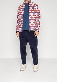 Kleurrijke ritssluitingenjack met geometrische patronen in rood, wit en blauw; gecombineerd met een marineblauw shirt en een donkerkatoenen broek; romige sneakers.