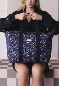 Kvinna håller en stor marinblå tygväska med blommönster och två handtag, sittande på en rutigt rosa och grå bänk.