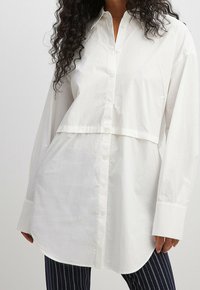 Chemise blanche à boutonner avec un design court, à manches longues et à ourlet incurvé ; en tissu coton lisse, avec un col classique et une texture subtile.