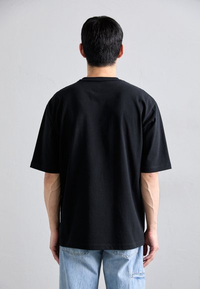 Man met kort zwart haar, gekleed in een simpele zwarte t-shirt en lichtblauwe jeans, staat tegen een eenvoudige lichtgrijze muur.