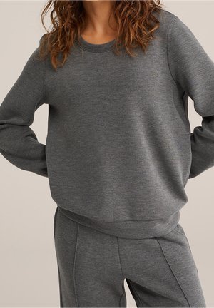 Personne portant un ensemble sweat-shirt et pantalon gris foncé assortis, mains sur les hanches, sur un fond neutre uni.