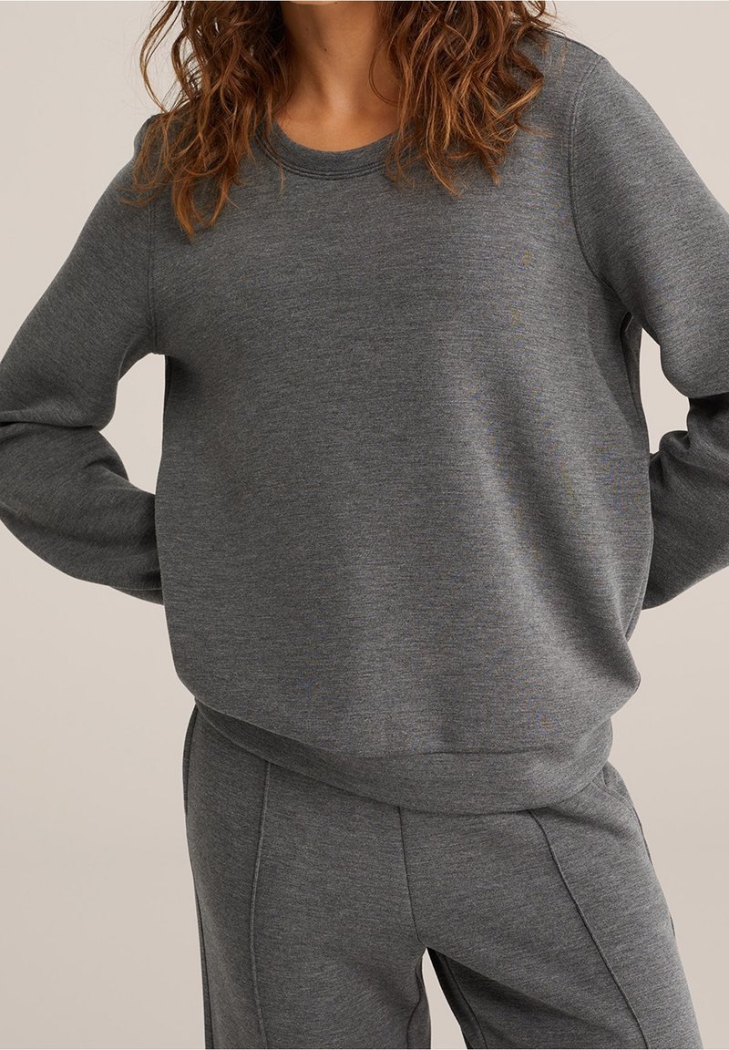 Personne portant un ensemble sweat-shirt et pantalon gris foncé assortis, mains sur les hanches, sur un fond neutre uni.