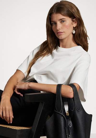 Camiseta branca de algodão com corte descontraído, combinada com calças pretas. Uma bolsa de tote em pele preta texturizada com alças duplas repousa no braço.