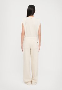 Beige sleeveless top with shoulder padding, paired with high-waisted beige wide-leg pants featuring back pockets and a small logo accent.