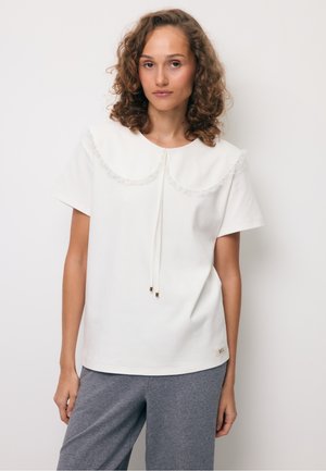 Młoda kobieta o kręconych włosach, ubrana w biały krótki top z dużym koronkowym kołnierzem i szare spodnie na jednolitym tle.