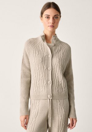 Junge Frau trägt eine beige kabelgestrickte, geknöpfte Strickjacke und passende Strickhosen vor einem schlichten Hintergrund.