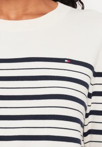 Hvit og marineblå stripete strikkegenser med ribbestruktur. Har en liten logo på venstre brystområde og en klassisk rund hals.