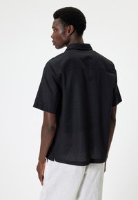 Camisa negra de manga corta en una tela ligera, con cuello y aberturas laterales para facilitar el movimiento. La parte trasera tiene un sutil detalle de costura.