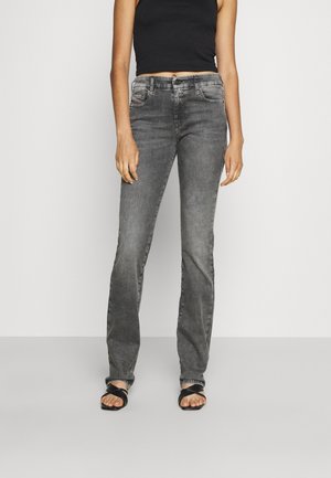 Kobieta ubrana w szare, wyblakłe jeansy z wysokim stanem, w połączeniu z czarnym tank topem i czarnymi sandałami z krzyżującymi się paskami na jednolitym tle.