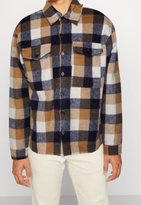 Flanellskjorta med rutmönster i brunt, marinblått och vitt. Har två bröstfickor, knäppning framtill och långa ärmar.