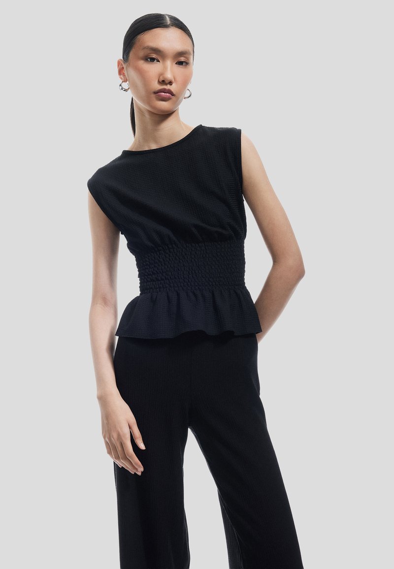 Top negro texturizado con una cintura fruncida ajustada y un dobladillo con volantes, que presenta tirantes anchos y una silueta relajada. Modelo contra un fondo gris.
