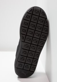 Semelle de chaussure noire avec des crampons à motif carré comprenant des formes d'étoiles et un logo Puma près du talon, inclinée contre une surface blanche.