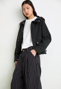 Sort cropped jakke med hætte, med sølv hardware og to frontlommer med lynlås, båret over en hvid t-shirt og sorte stribede bukser.