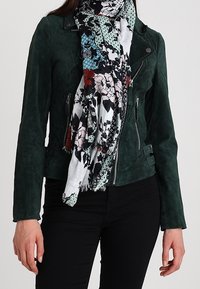 Veste biker en daim vert foncé avec des fermetures éclair et des accents de panneaux, associée à une écharpe blanche et noire à motif floral. Pantalon noir.