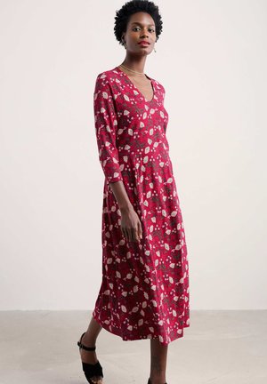 Robe florale rouge à manches trois-quarts, décolleté en V, longueur genoux, fabriquée dans un tissu léger, présentant un motif de fleurs blanches et vertes.