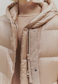 Beige padded jacket with a hood, featuring a shiny exterior, visible zippers, and contrasting fabric for the inner layer. Textured knit collar.
