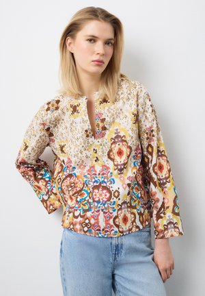 Mujer joven con cabello rubio, vistiendo una blusa de manga larga con estampado colorido y jeans azul claro, de pie contra un fondo blanco.