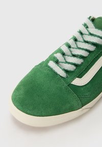 Baskets en daim vert avec une bande diagonale blanche, une semelle blanche et des lacets en coton blanc sur un fond blanc uni.