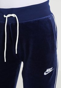 Námořnické modré velurové tepláky s bílým šňůrkou a bočními pruhy, na levém stehně s bílým logem Nike.