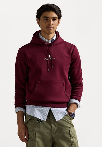 Felpa maroon con tasca a marsupio, cappuccio con cordino e logo ricamato. Indossata sopra una camicia a righe blu chiaro e pantaloni cargo beige.