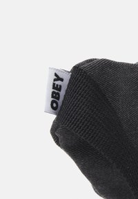 Μαύρος υφασμάτινος ιμάντας με υφή, που διαθέτει μια λευκή ραμμένη ετικέτα που γράφει "OBEY". Ο ιμάντας έχει λείο, στρογγυλεμένο σχήμα.