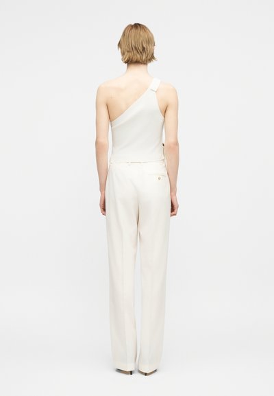 Άτομο που φοράει λευκό τοπ ενός ώμου και λευκό φαρδύ παντελόνι ψηλόμεσο, στέκεται με την πλάτη γυρισμένη σε απλό ανοιχτό φόντο.