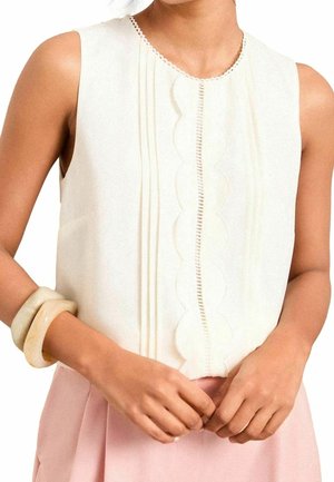 Femme portant un haut blanc sans manches avec des détails festonnés et plissés à l'avant, une jupe rose clair et deux grands bracelets crème à son poignet.