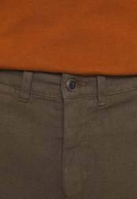 Hnědé kalhoty z tkané látky s texturovaným povrchem, předním zapínáním na knoflík a kontrastním opáleným prošíváním. Pas je mírně elastický.