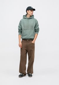 Zelená mikina s kapucňou a predným vreckom, doplnená voľnými hnedými nohavicami. Čierne tenisky dotvárajú ležérny outfit.