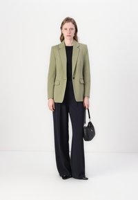 Olivový zelený blejzer na mieru s jedným gombíkom, spárovaný s čiernym topom a námorníckymi širokými nohavicami. Model drží malú čiernu kabelku.