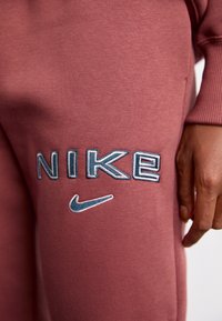 Nike sweatpants i en dammig rosafärg, med blå broderad "NIKE"-text och en liten blå swoosh-logotyp. Slät textur, avslappnad design.