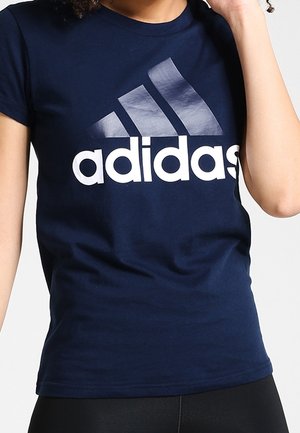Osoba ubrana w granatową koszulkę Adidas z dużym białym logo Adidas na klatce piersiowej.