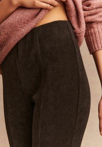 Közelkép egy személyről, aki magas derekú sötétbarna bordázott leggingset és egy rózsaszín kötött pulóvert visel, amelyet felemeltek, hogy a derékpánt látható legyen.