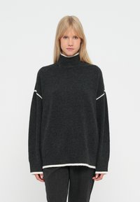 Tumši pelēks adīts turtlenecks džemperis ar baltām akcentiem ap kaklu un apakšu. Iekļauj kritušos plecus un brīvu piegriezumu.
