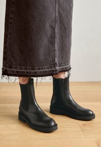 Bottes noires en cuir à la cheville avec une semelle épaisse, des panneaux élastiques sur le côté et des fermetures éclair ; portées avec une jupe sombre effilochée sur un sol en bois.