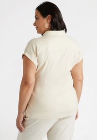 Camicia corta beige chiaro a maniche corte con colletto, polsini arrotolati e sottili decorazioni sulle spalle, caratterizzata da una vestibilità rilassata e dettagli sulla cucitura posteriore.