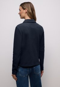 Femme aux cheveux châtain clair portant une veste texturée bleu marine foncé et un jean bleu, vue de dos sur un fond uni gris clair.