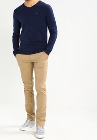 Man som bär en marinblå långärmad tröja, beige chinos och grå sneakers, stående med händerna i fickorna mot vit bakgrund.