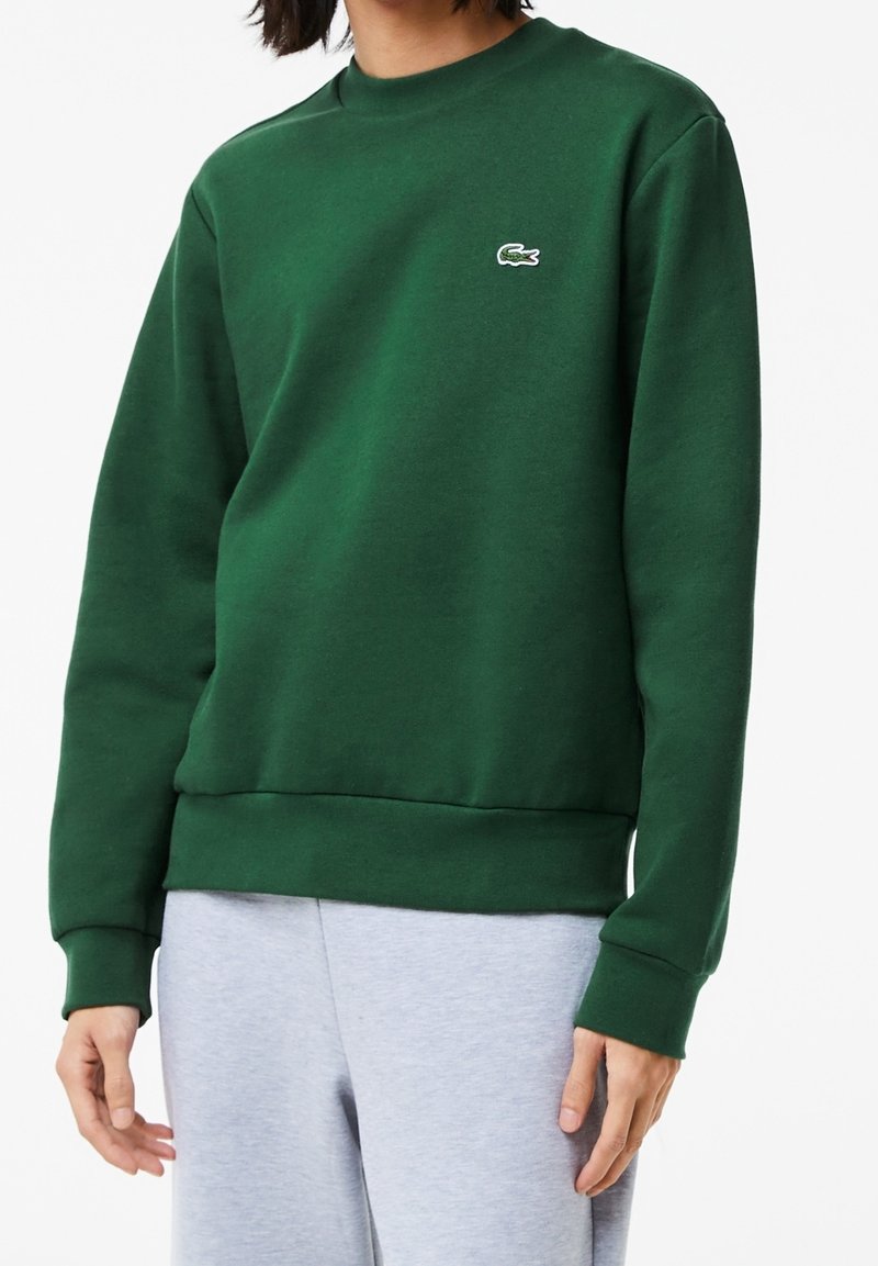 Personne portant un sweat-shirt à manches longues vert foncé avec un petit logo crocodile blanc sur la poitrine gauche et un pantalon gris clair.